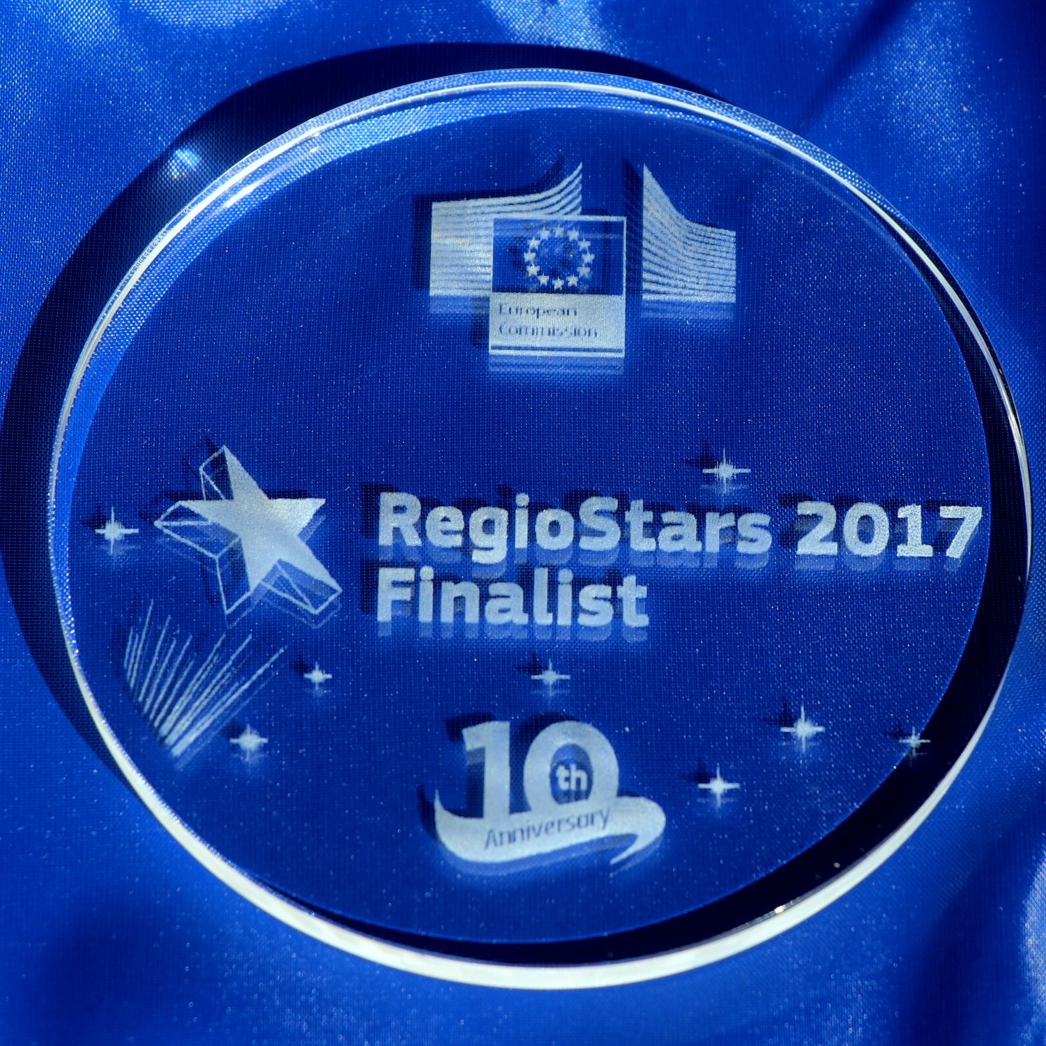 Nagroda w postaci płaskiego okręgu ze szkła z grawerowanymi kształtami gwiazd, flagą Unii Europejskiej i napisem w języku angielskim RegioStars 2017 Finalist.