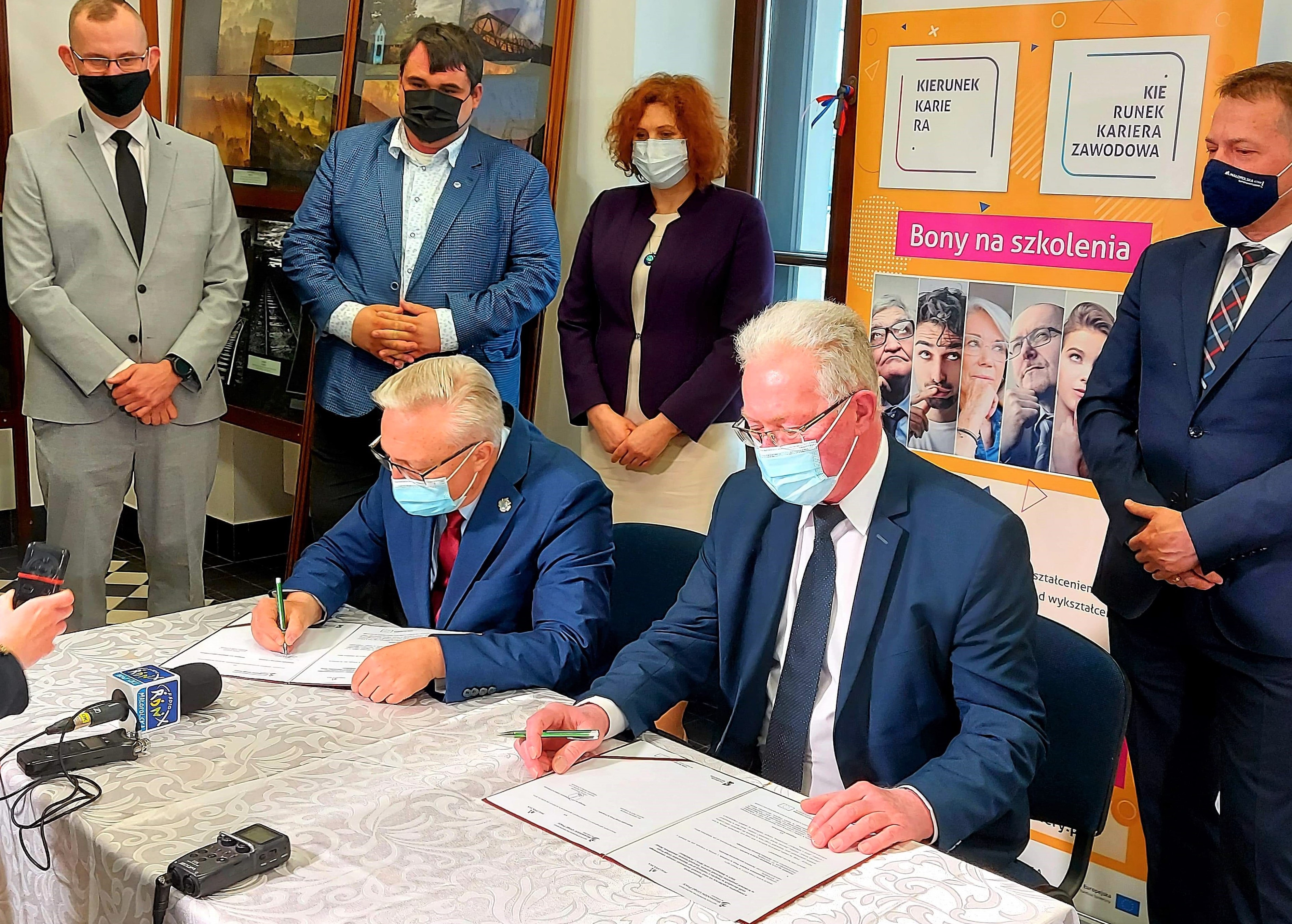 na zdjęciu są osoby które w ręce trzymają długopis i podpisują dokument-porozumienie, siedzą przy stole, po lewej stronie siedzi starszy szpakowaty mężczyzna w okularach, ubrany  jest w garnitur i czerwony krawat, a po prawej stronie siedzi też mężczyzna szpakowaty w okularach, ubrany w garnitur i granatowy  krawat.