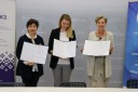 Trzy kobiety stojące obok siebie, każda trzyma w rękach otwartą teczkę z podpisanym dokumentem.