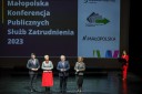 Na scenie Opery Krakowskiej obok siebie stoją 2 kobiety i 2 mężczyzn. W tle za nimi wyświetlona nazwa konferencji, z boku stoi prowadząca konferencję prezenterka.