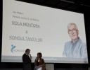 Dwie osoby rozmawiają ze sobą na scenie, stojąc. W tle ekran, na którym jest widoczna sylwetka prelegenta: Jana Mądrego i tytuł jego wystąpienia: Rola mentora a konsultanta HR.