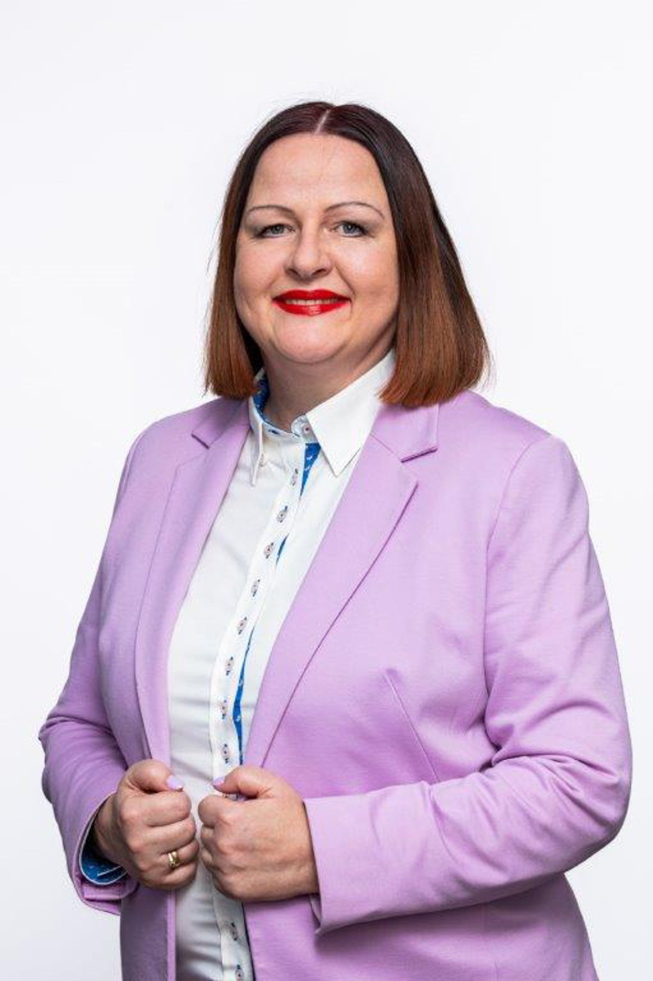 Kobieta ubrana w jasnofioletową marynarkę i białą koszulę, z prostymi włosami do ramion. Obiema rękami trzyma krawędzie marynarki.