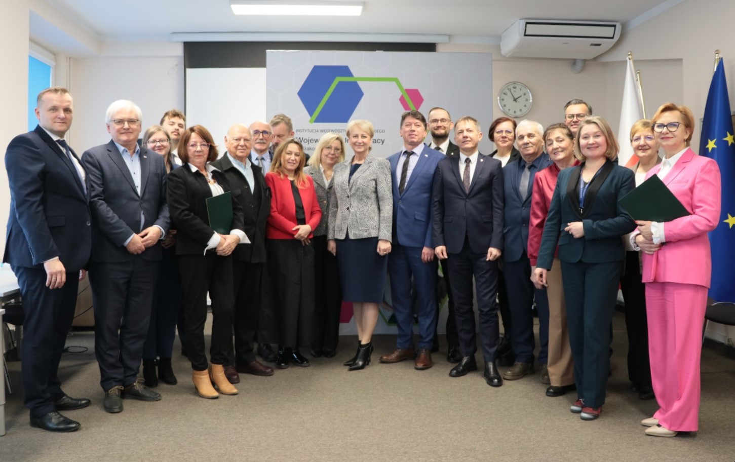 Grupa około 20 dorosłych osób w strojach formalnych pozuje do wspólnego zdjęcia w sali konferencyjnej, z flagami i banerem w tle.