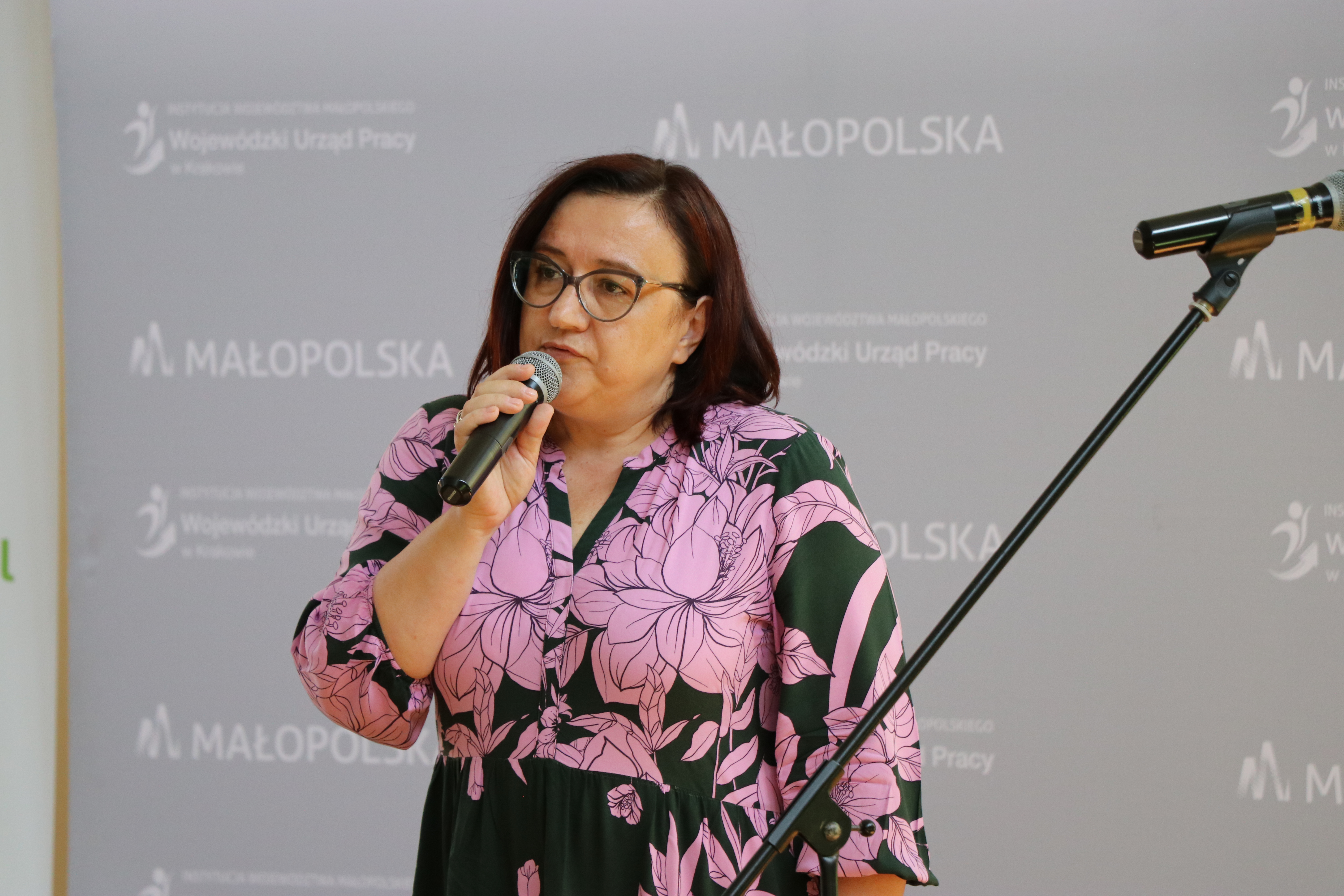 Zdjęcie przedstawia kobietę o brązowych włosach, mówiącą do mikrofonu. W tle napis Małopolska oraz Wojewódzki Urząd Pracy w Krakowie.