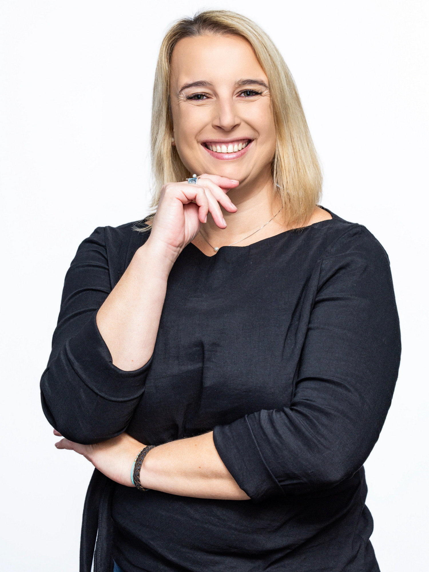 Uśmiechnięta kobieta, blondynka z prostymi włosami opadającymi na ramiona. Prawą dłoń opiera o podbródek.