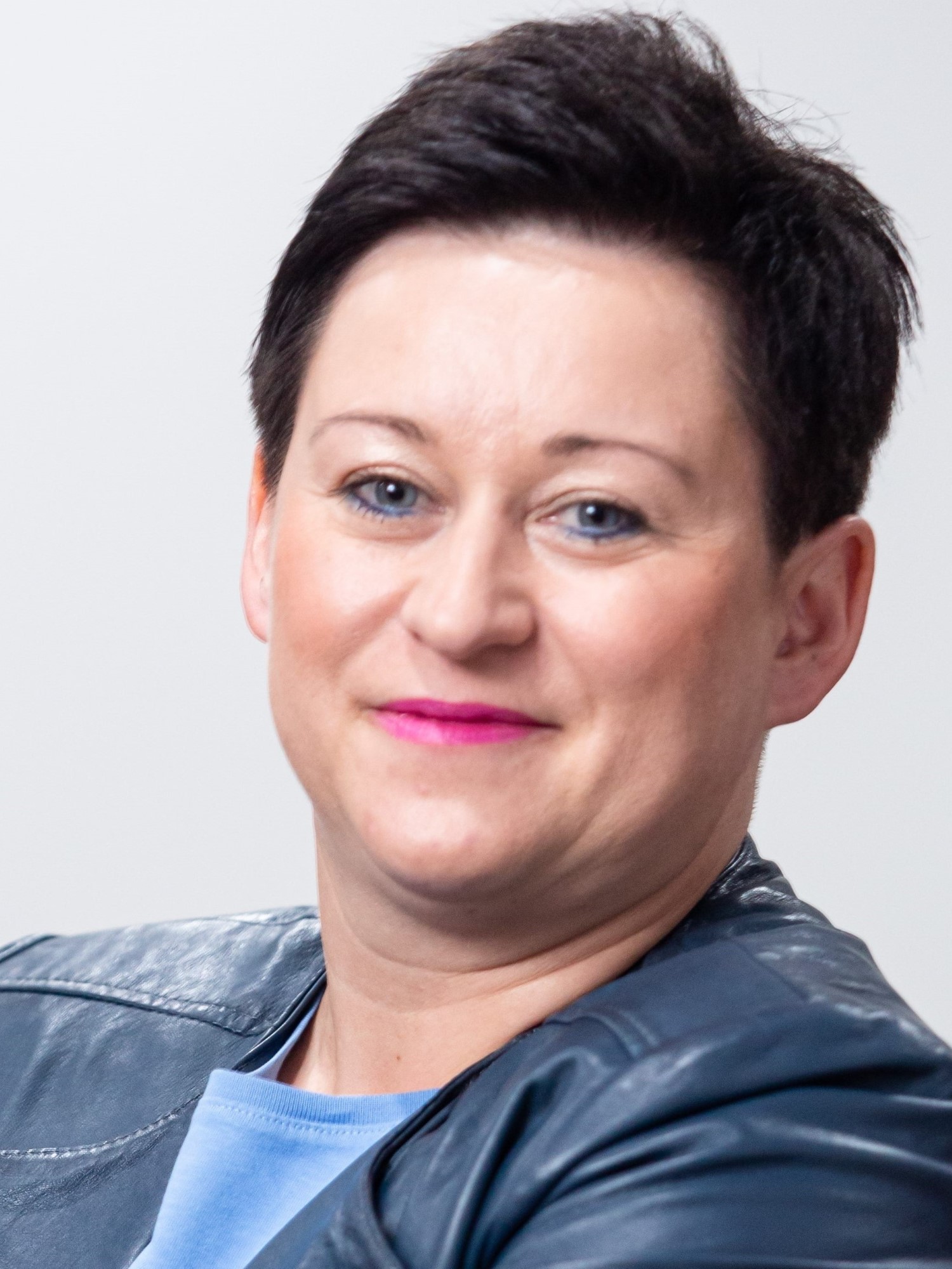 Uśmiechnięta kobieta z krótkimi ciemnymi włosami. Ubrana w ciemną skórzaną marynarkę, pod którą ma jasnoniebieski top.