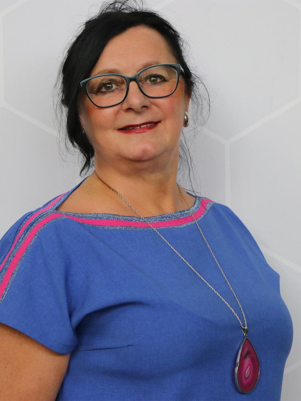 Uśmiechnięta ciemnowłosa kobieta w okularach, ubrana w niebieski strój z różowymi akcentami.