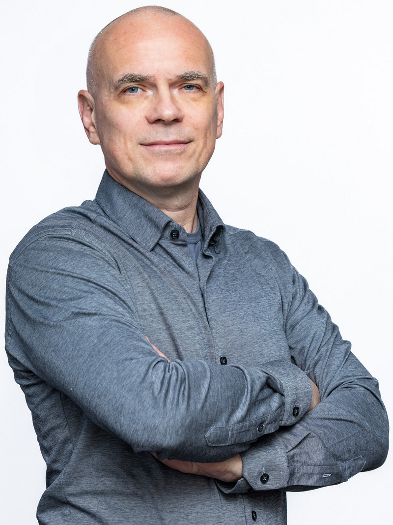 Lekko uśmiechnięty mężczyzna, łysy, stoi z założonymi na siebie rękami. Ubrany w szarą koszulę z długim rękawem, zapinaną na guziki.