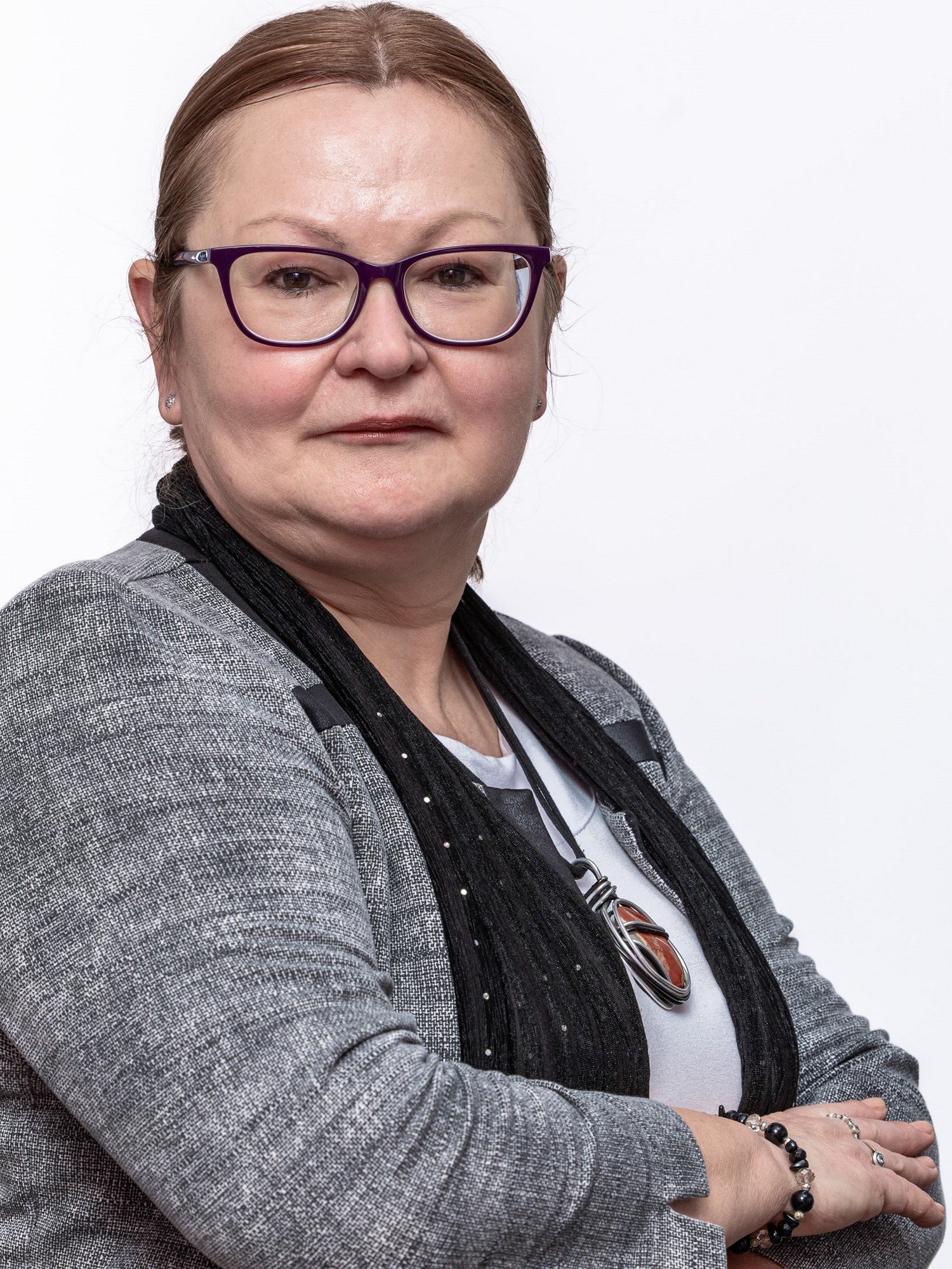 Kobieta w stylowych okularach, włosy ma zaczesane i związane z tyłu. Ubrana w stonowaną bluzę i jasny top. Stoi zwrócona bokiem z rękoma założonymi na siebie.