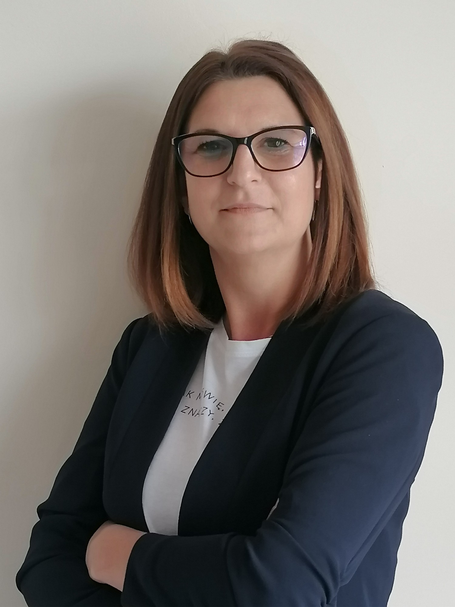 Kobieta w okularach, z włosami opadającymi na ramiona. Ubrana w ciemną marynarkę, pod którą widać białą bluzkę. Ręce założyła na siebie, stoi lekko odwrócona bokiem.
