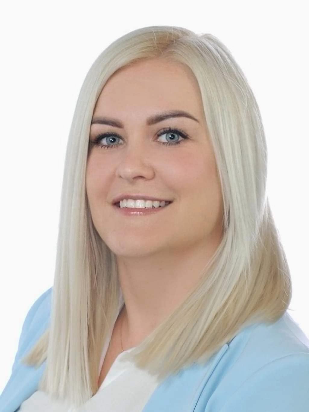 Uśmiechnięta platynowa blondynka, z prostymi włosami opadającymi na ramiona. Ubrana w białą bluzkę i jasnoniebieski żakiet.